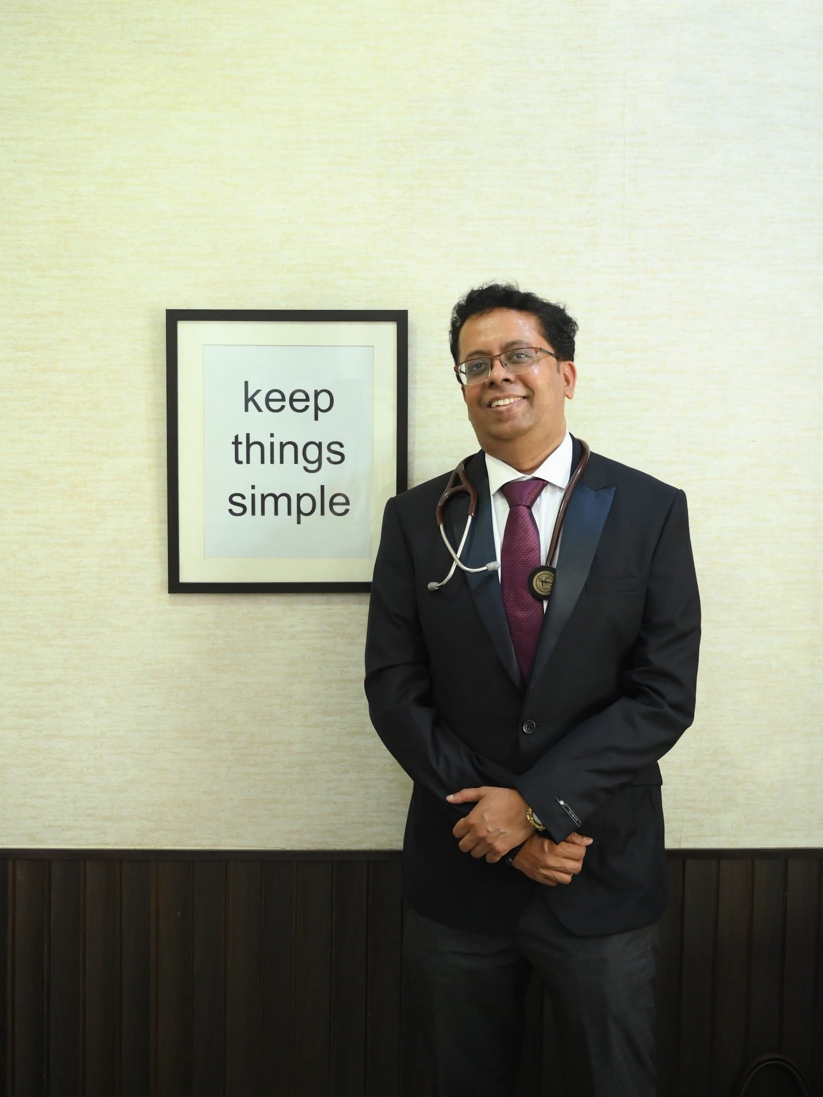 Dr. Ajay Kaduskar standing beside a framed 'keep things simple' print in his consulting room